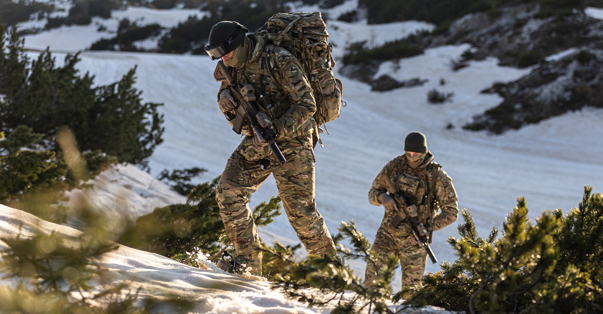 Two fully equipped operators in winter tactical clothing ascending a snowy hillside on a sunny winter day.